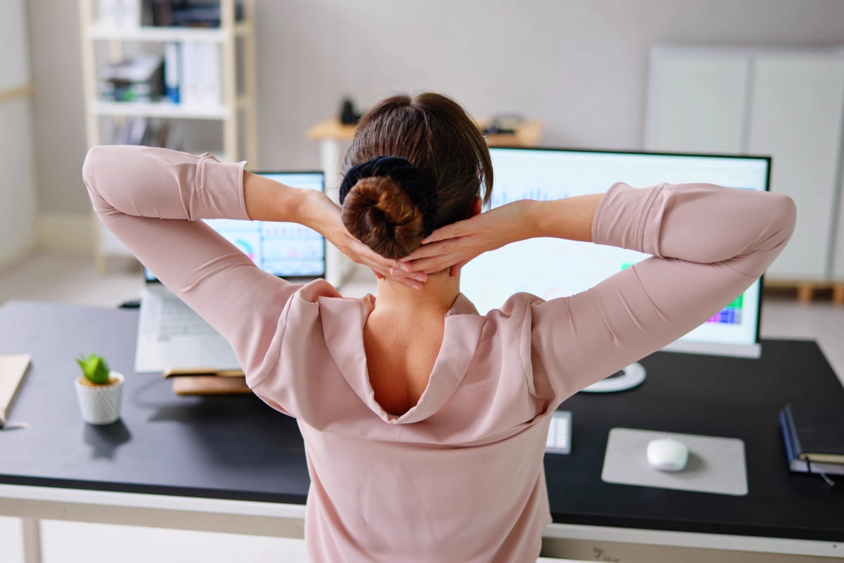 Person stretching at a workstation in front of dual monitors to reduce neck and back strain during the workday.
