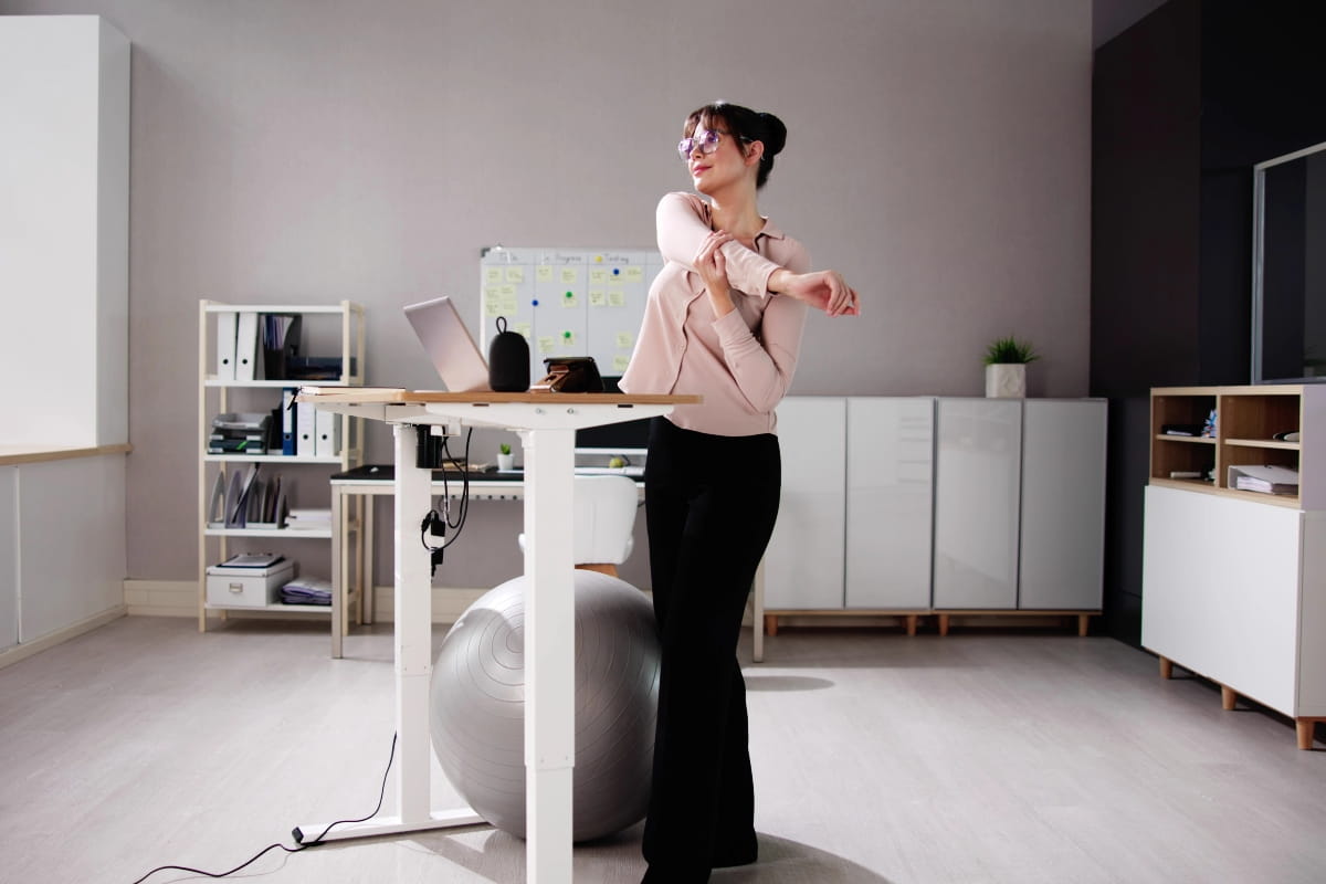 Standing Desk Posture: How to Stand Comfortably All Day 3 Office worker taking a stretch break beside an adjustable desk and exercise ball.