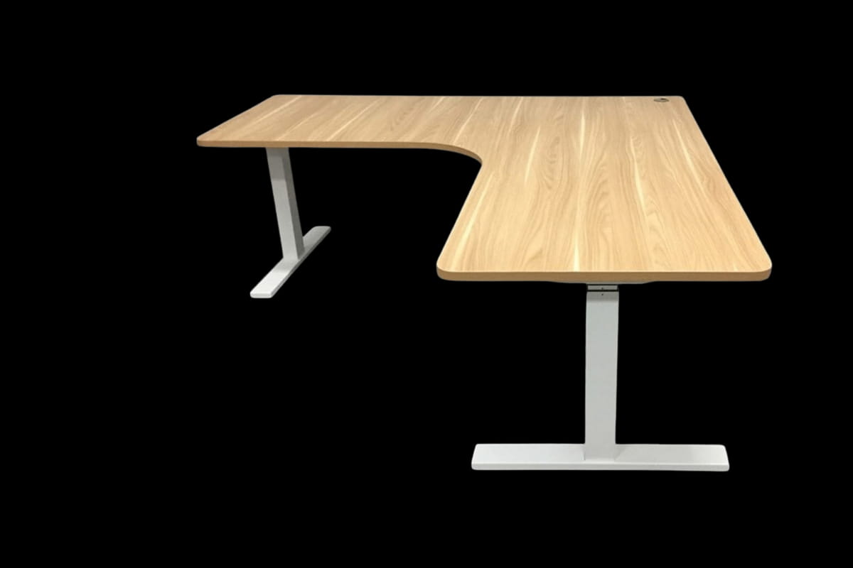 L-shaped standing desk with a light wood desktop, curved inner corner and silver metal legs against a black background.