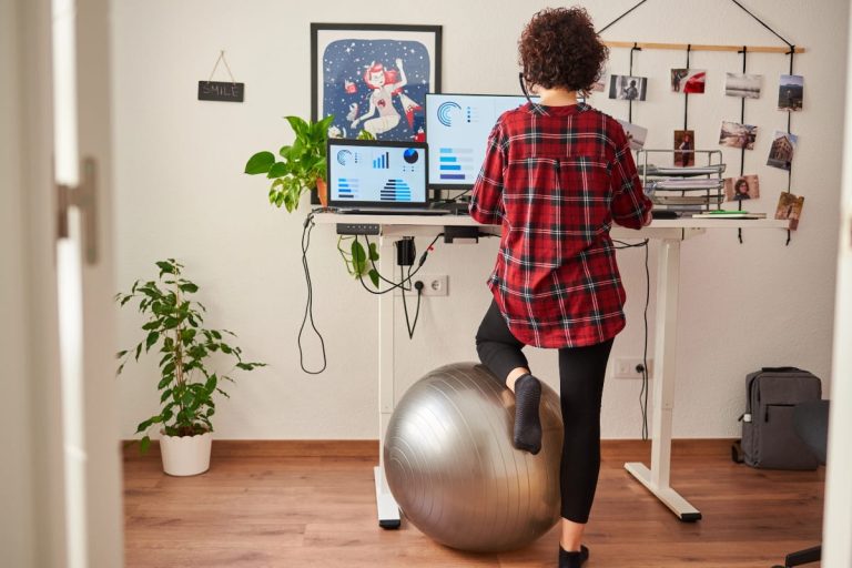 Standing desk workstation with a person alternating posture using a balance ball under one leg, highlighting the ergonomic debate about are standing desks good for you.