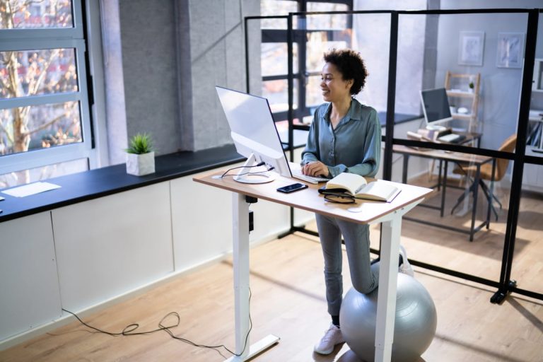 Employee working at a raised desk with a balance ball nearby, showing how to transition to a standing desk gradually.