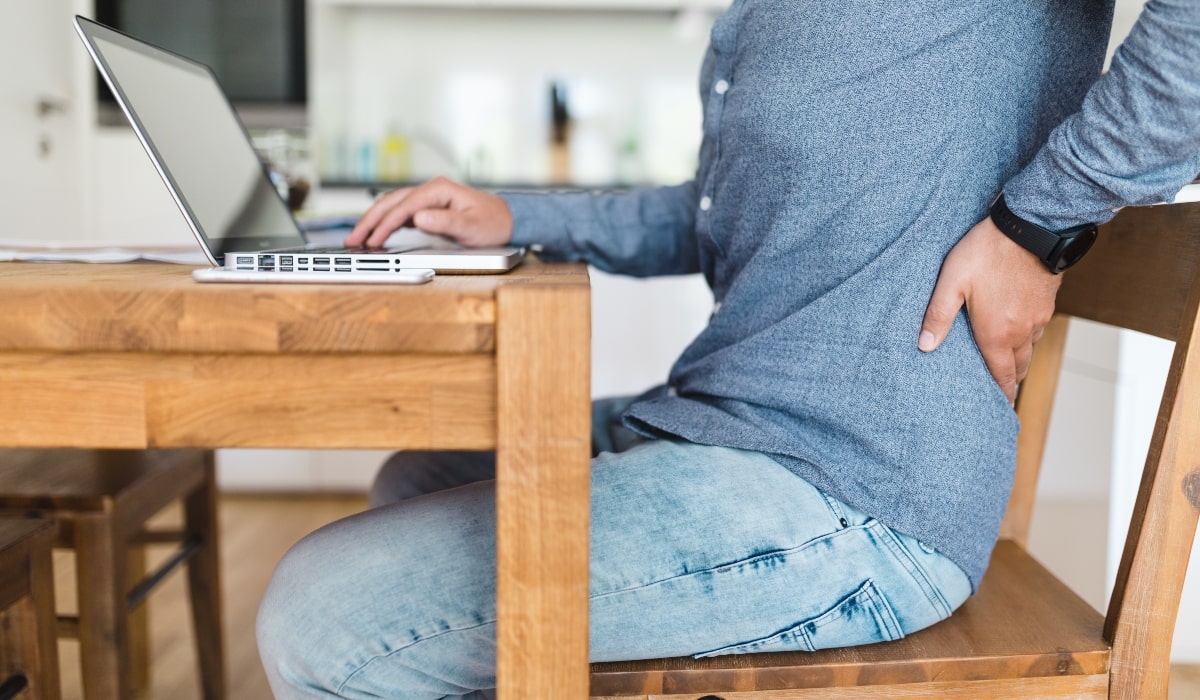 A man working from home at his dining table, with his hand on his back due to lower back pain.