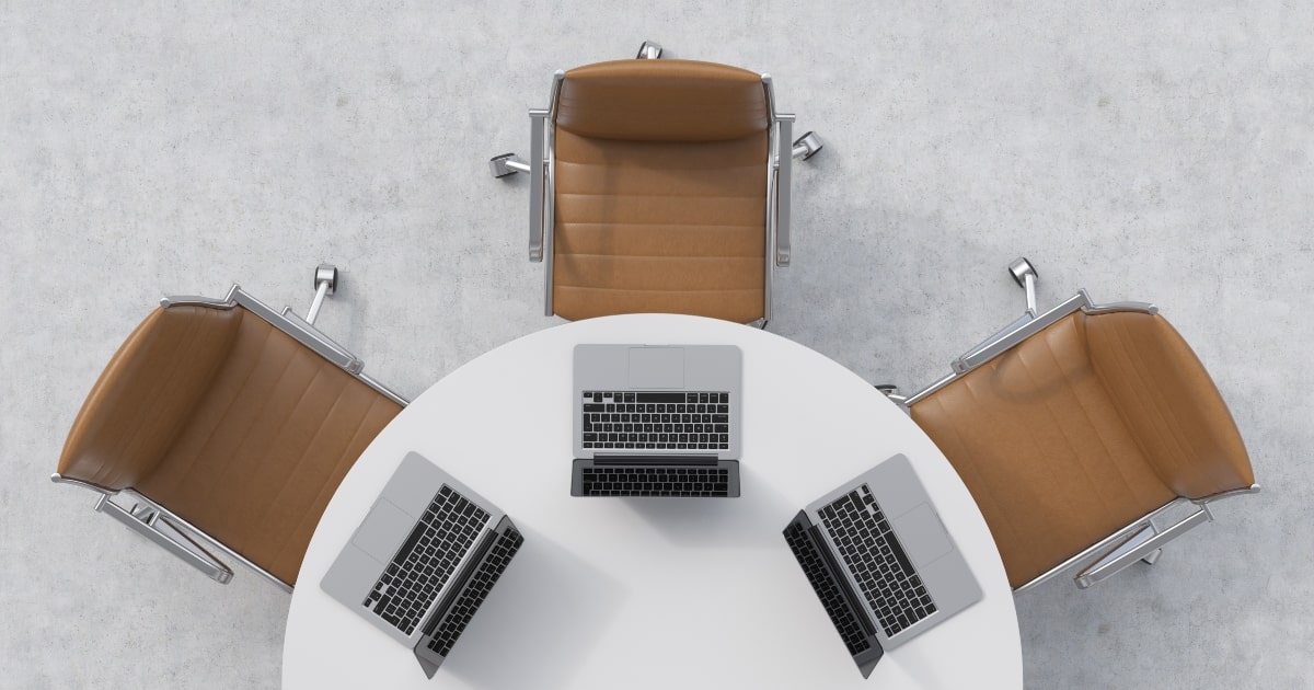 Top view of a white round table, three brown leather chairs and three laptops.