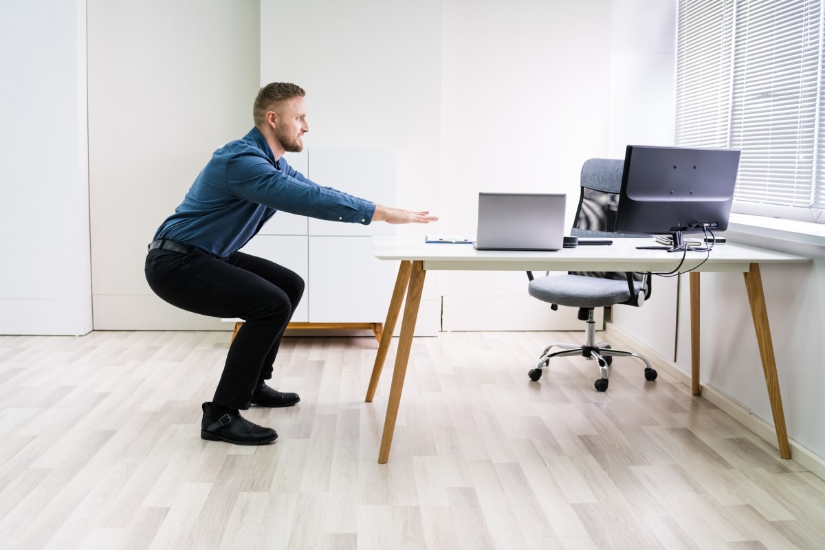 A man in business attire performing chair squats as part of his office chair exercises routine. 