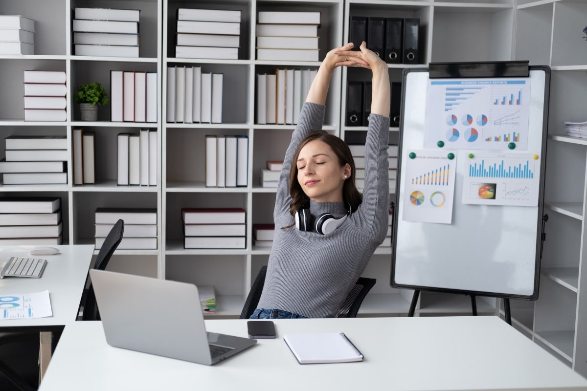 A woman sitting at her desk stretching with her arms extended above her head, taking advantage of small movement breaks throughout her workday to promote physical and mental wellness. 