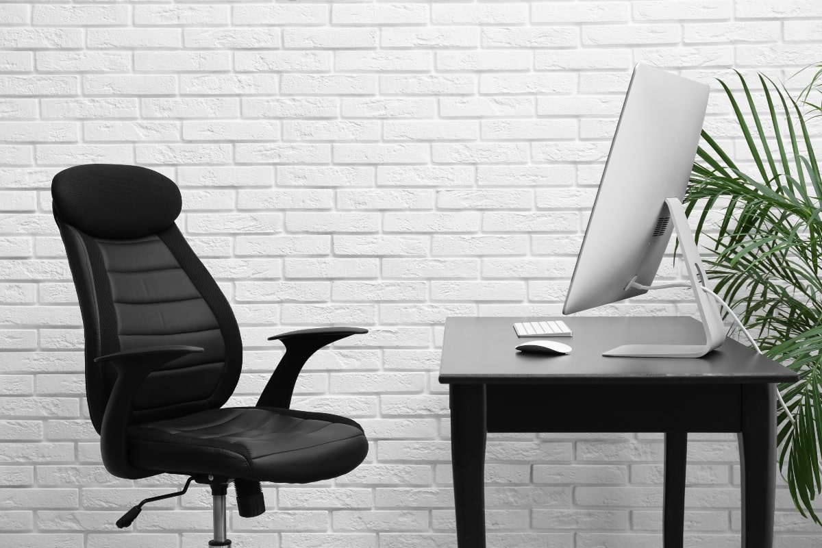 An ergonomic leather office chair positioned in front of a minimalist desk, against a backdrop of a white brick wall, showcasing the durability and comfort that leather brings to executive office settings.
